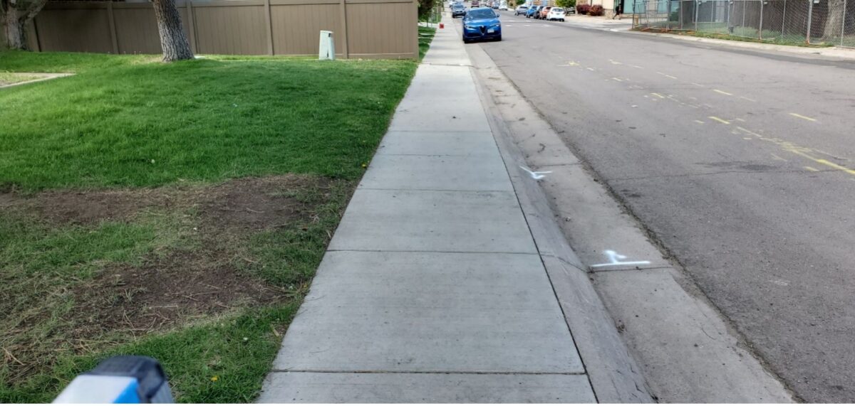 A photograph of the right-of-way outside of a Denver day care. Grass is on the left, sidewalk is in the center, and the street is on the right. Parked cars, a fence, and a tree are visible in the background.