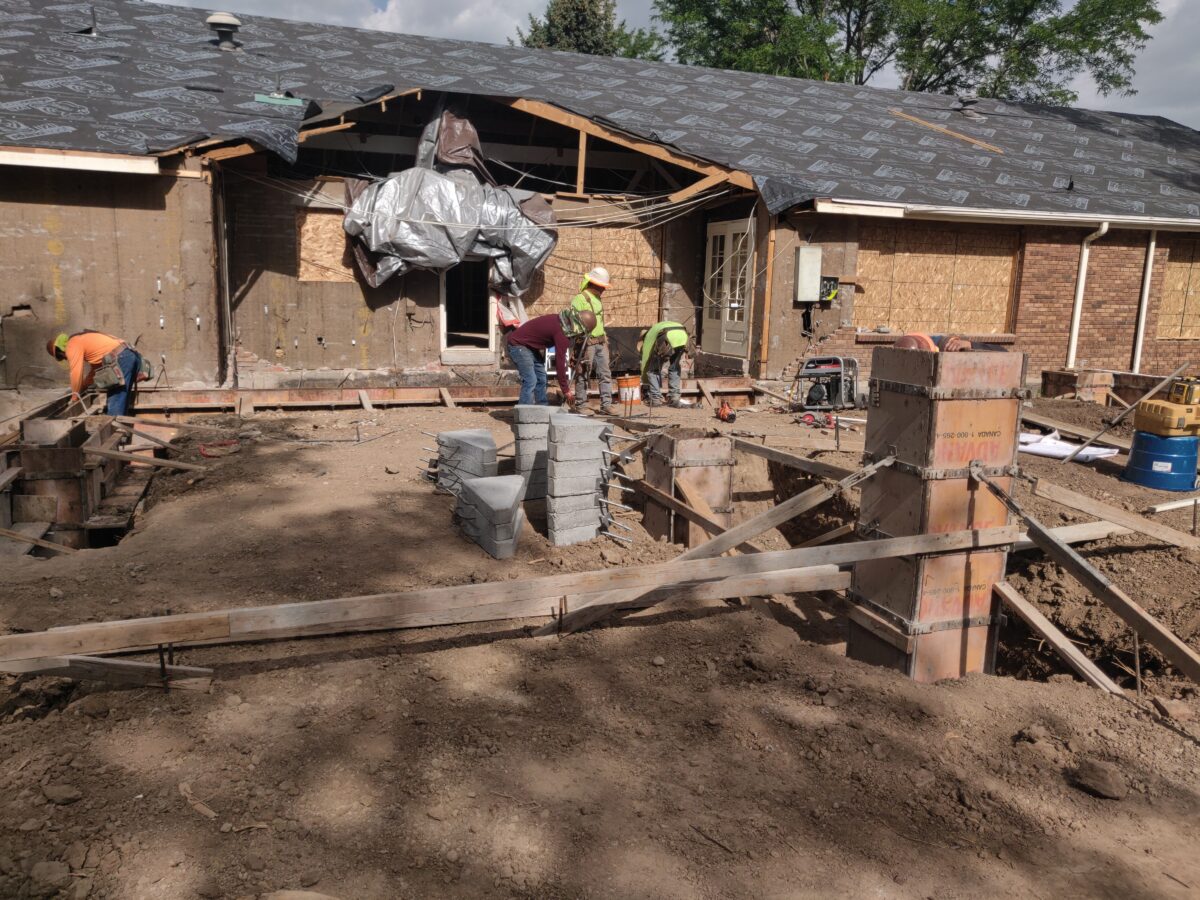 An image of construction workers working on a home remodel project. Exposed lumber, dirt, and bricks dominate the photo.