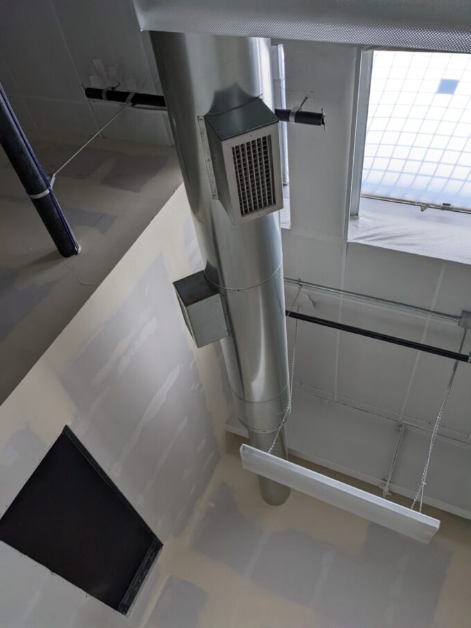 A photo taken from below of smooth, silver metal exposed ductwork. The room has white walls and ceilings that look like they are in the process of being completed, and a skylight in the top right corner.