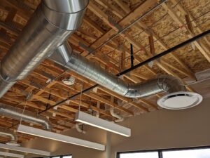 A photograph taken from below shows silver spiral exposed ductwork is shown against an exposed plywood and wooden beam ceiling with lights hanging from it. The room has cream colored walls.