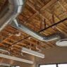 A photograph taken from below shows silver spiral exposed ductwork is shown against an exposed plywood and wooden beam ceiling with lights hanging from it. The room has cream colored walls.