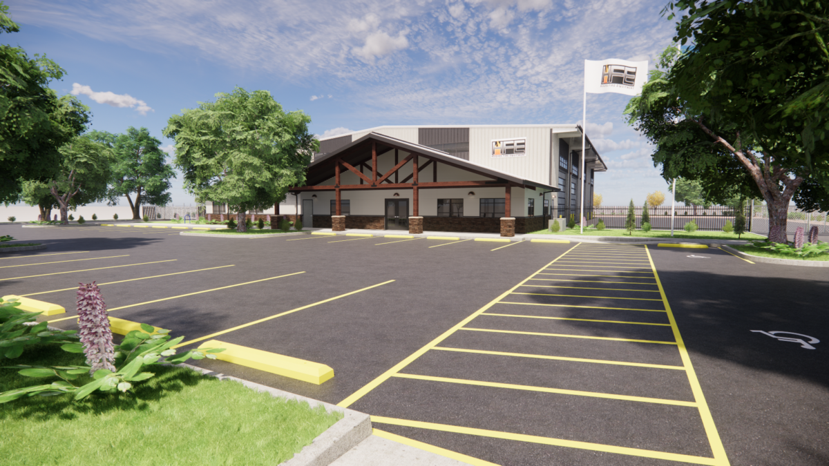 An example of virtual reality modeling of the exterior of a building. There is a lawn with a flowering plant in the foreground on the left. The center of the image is a parking lot. The building is centered in the background, with trees on both sides, as well as a flag with the Hughes Fire Equipment logo to the right. Behind the flag, there is a fence. Behind the fence, there are three fire truck bays.