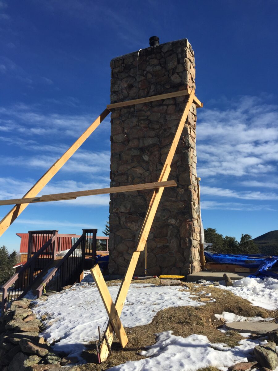 A photograph of a fireplace freestanding aside from some wooden supports. The fireplace is in the midst of a construction site, and a blue sky is prominent in the background. During the building process, the owners wanted to keep this fireplace and rebuild the home around it.