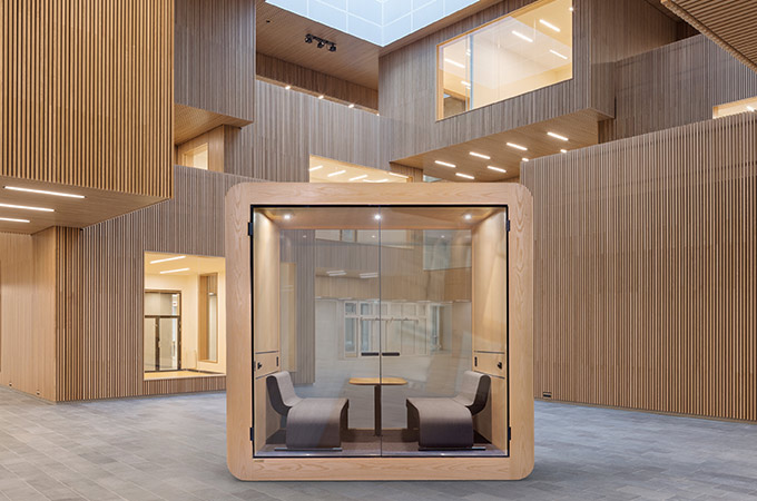 An example of an acoustic pod. It is a small standalone structure in the middle of a larger room that has large glass French doors, two chairs with a table in between, and soft lighting inside.