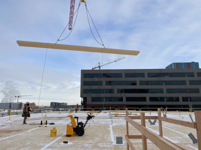 A photo shows a crane (mostly off-screen) hoisting a cross-laminated timber (CLT) panel being placed on top of construction at T3. It appears to be a somewhat cloudy winter day, with light snow on the ground.