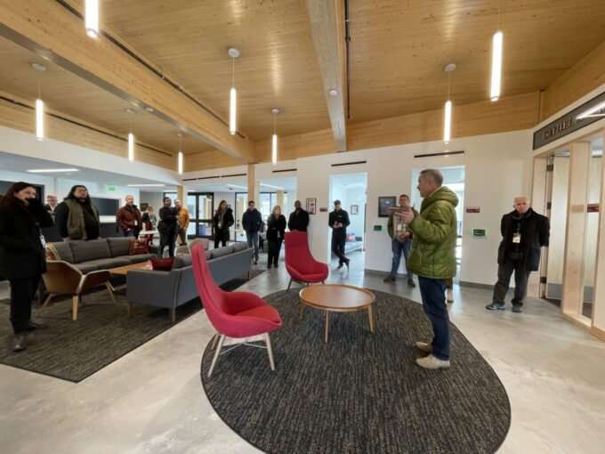 A group of people is shown inside the Burwell Center. A white man in a green puffy coat appears to be speaking to the group.