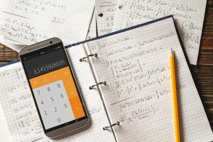 An Adobe stock image: Solving mathematical problems in a notebook . Phone with calculator app on wooden desk.