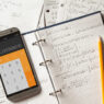 An Adobe stock image: Solving mathematical problems in a notebook . Phone with calculator app on wooden desk.