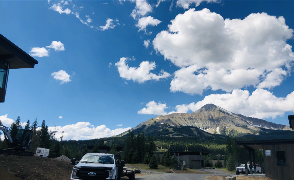 A photograph of mountains as seen from Big Sky, MT with a white pickup truck in the lower left corner, parked and facing the camera.