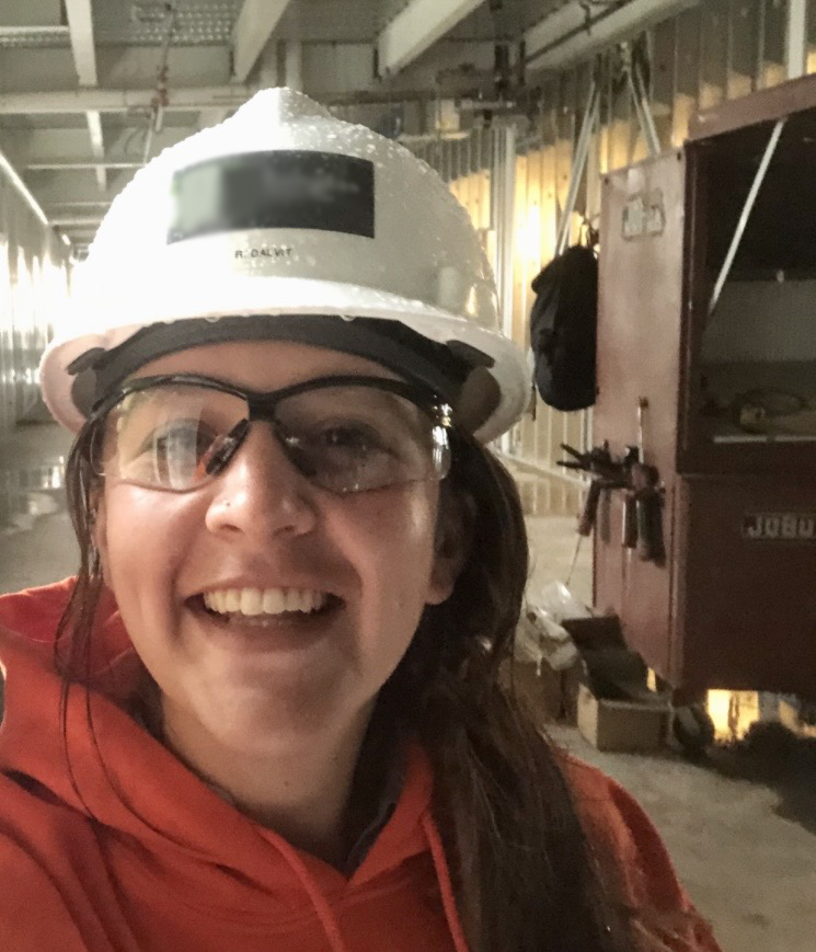 A selfie of the author. She is smiling, wearing a white hard hat and clear safety glasses, with a red hoodie on and her hair pulled back and over one shoulder. She is on an active construction site.