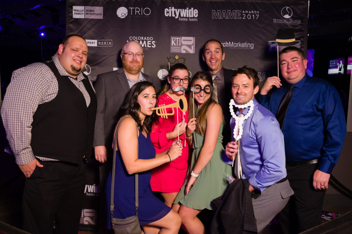 A group of people pose in formal clothes with photo booth props at an event.