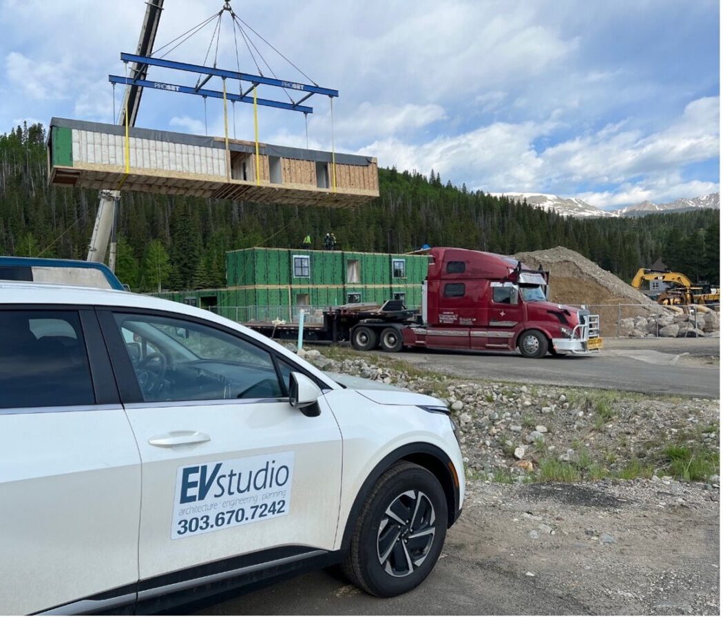 EVstudio's white company vehicle is shown in the foreground of the construction site for the Justice Center Apartments in Breckenridge, CO. Modular construction of volumetric design is shown in the background.