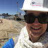 A selfie of Katelyn at a construction site. She is wearing a blue shirt, a white scarf with polka dots, black sunglasses, and a white hardhat. The hardhat has a sticker that has the EVstudio logo with Where Girls Rule! in hot pink text underneath.