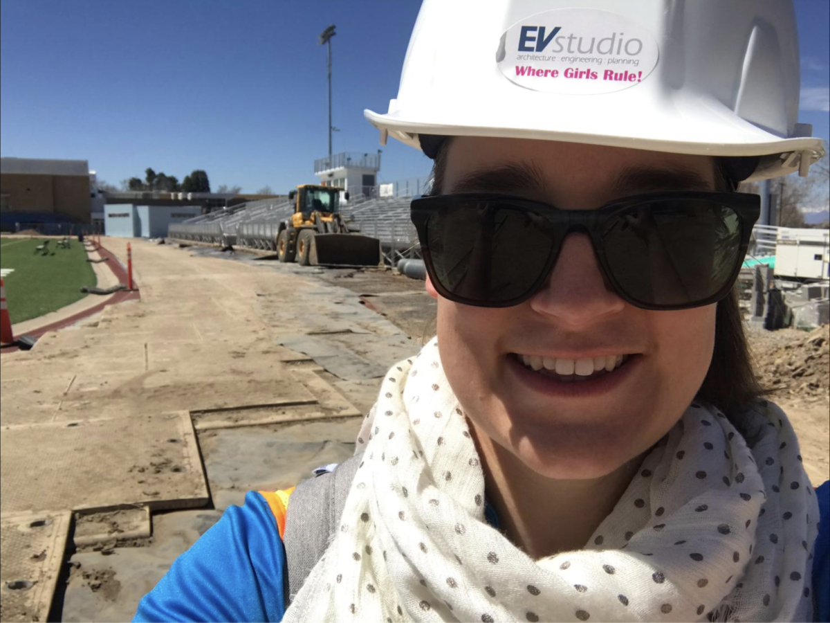 A selfie of Katelyn at a construction site. She is wearing a blue shirt, a white scarf with polka dots, black sunglasses, and a white hardhat. The hardhat has a sticker that has the EVstudio logo with Where Girls Rule! in hot pink text underneath.